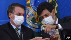 Mengenakan masker Menteri Kesehatan Brazil Luiz Henrique Mandetta (kanan), membubuhkan penyanitasi tangan ke tangan Presiden Jair Bolsonaro saat berlangsungnya konferensi pers di Istana Presiden Planalto di Brasilia, Brazil, 18 Maret 2020. (Foto: dok).