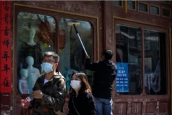 In this April 12, 2020, photo, a Chinese couple walks past a worker wiping window panels at a clothing shop in Beijing.