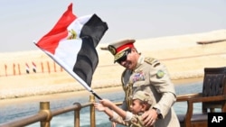 In this picture provided by the office of the Egyptian Presidency, President el-Sissi smiles at a boy dressed in a tiny military uniform as he waves the national flag from a monarchy-era yacht that sailed to the venue of a ceremony for extension of the Su
