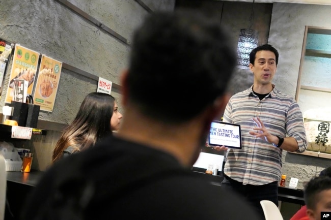 Frank Striegl, right, a guide of Tokyo Ramen Tours, explains foreign participants of a ramen tasting tour at Shinbusakiya, a ramen shop which offers "Hokkaido classics," at Shibuya district on April 2, 2024, in Tokyo. (AP Photo/Eugene Hoshiko)