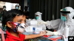 Seorang anak laki-laki mengenakan masker, menunggu disuntik menunggu petugas kesehatan melakukan tes antibodi virus corona di sebuah desa di Bali, Rabu, 27 Mei 2020. (AP Photo / Firdia Lisnawati)