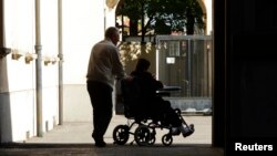 An elderly man pushes a woman in her wheelchair. 