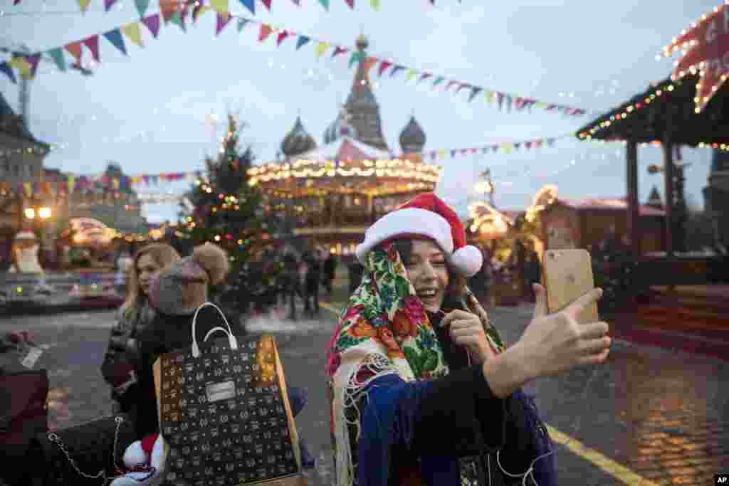 A woman dressed in Russian traditional headscarf and Santa Claus hat talks by phone video call at the Christmas Market in Red Square, with St. Basil's Cathedral in the background, in Moscow, Russia.