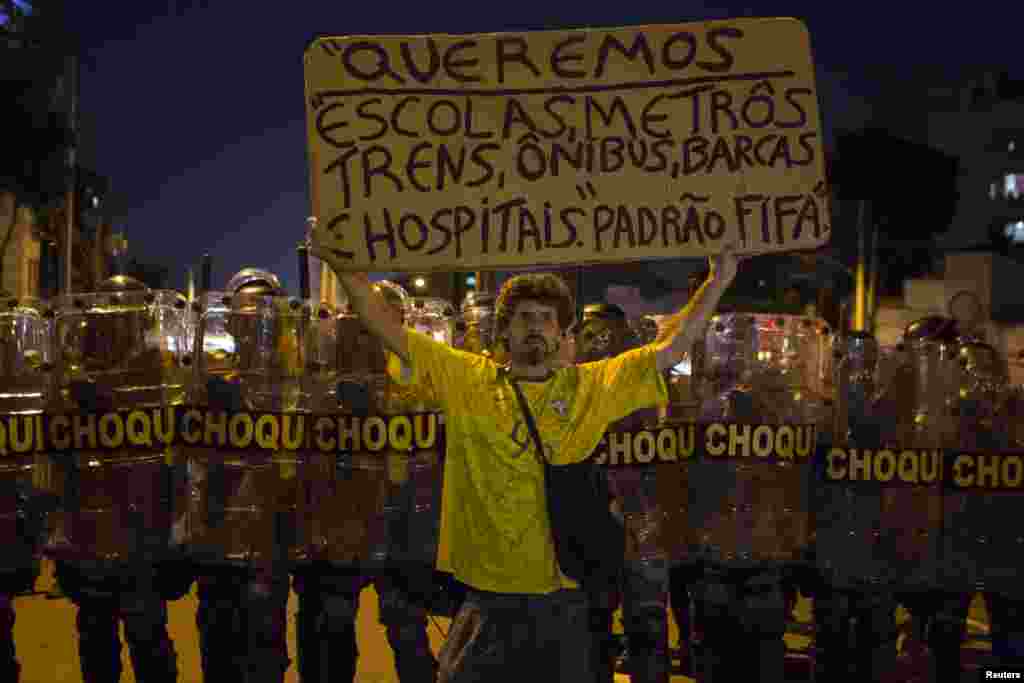 A protester holds a sign that reads &quot;We want schools, subways, trains, buses, boats, hospitals of FIFA standard&quot; in front of riot police during a demonstration against the public spending for the 2014 World Cup, in Rio de Janeiro, June 15, 2014.