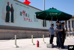 FILE - A man looks at his smartphone next to Chinese policemen and a paramilitary policeman standing guard near a giant electronic screen showing a Chinese government propaganda message reading "Dream road ahead, People's Communist party" on Tiananmen Square in Beijing, China on May 28, 2014.