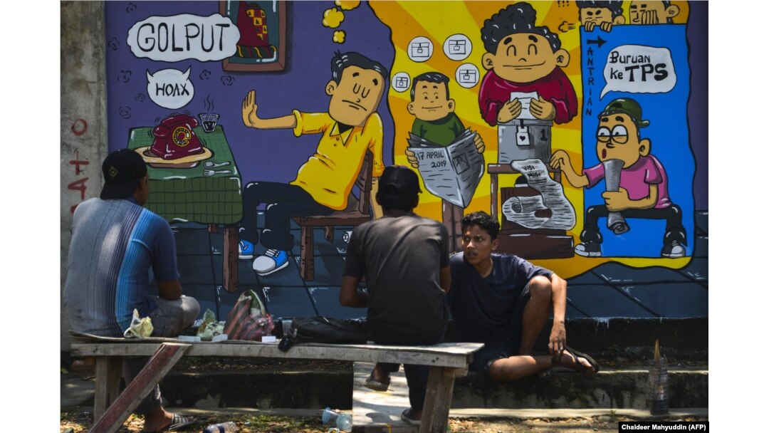 Orang-orang duduk di depan mural kampanye pemilihan umum Indonesia menjelang pemilihan bulan depan di Banda Aceh, Aceh pada 17 Maret 2019. (Foto: AFP/Chaideer Mahyuddin)