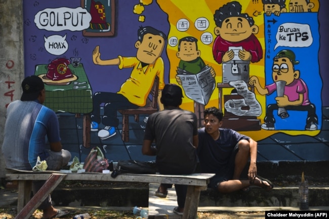 Orang-orang duduk di depan mural kampanye pemilihan umum Indonesia menjelang pemilihan bulan depan di Banda Aceh, Aceh pada 17 Maret 2019. (Foto: AFP/Chaideer Mahyuddin)