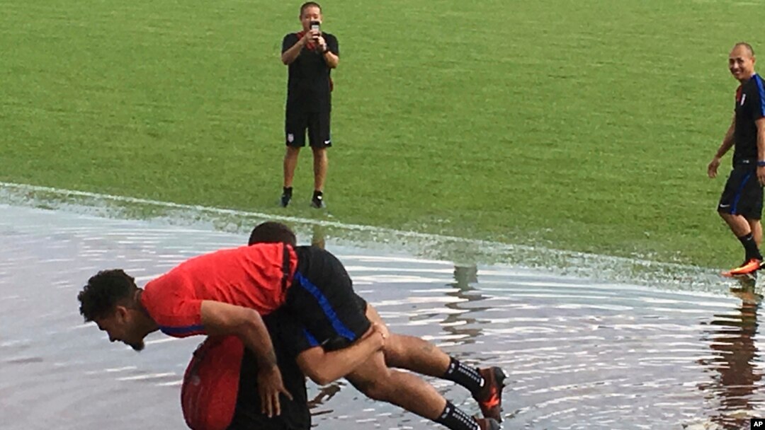 Pemain tim nasional AS, DeAndre Yedlin digendong pelatih Luis Ramirez melewati genangan air di sekitar lapangan tempat latihan, di Couva, Trinidad, Senin, 9 Oktober 2017. 