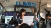  Passengers listen as El Bus TV’s Claudia Lizardo reads a news report. Holding the frame is co-founder Laura Castillo. The service began in May in Caracas, Venezuela, and has spread to other communities. (C. Alcalde/VOA Spanish Service) 