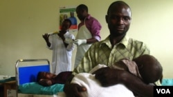 Savio Niwagaba holds his newborn baby as his wife Chrisente, behind him, has a contraceptive implant inserted in her arm, Kanungu, Uganda, June 19, 2012. (VOA / Hilary Heuler)