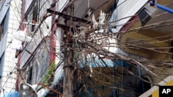 A woman, top left, stands at a balcony of her apartment near a lamp post jumbled with electric cables in Rangoon, Burma, Jan. 27, 2014.