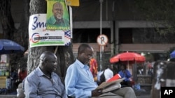 Tanzanians sit next to a tree, underneath an election poster for ruling party presidential candidate John Magufuli, as they await election results in Dar es Salaam, Tanzania, Oct. 27, 2015.