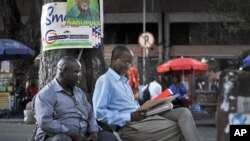 FILE - Tanzanians sit next to a tree, underneath an election poster for ruling party presidential candidate John Magufuli, as they await election results in Dar es Salaam, Tanzania, Oct. 27, 2015.