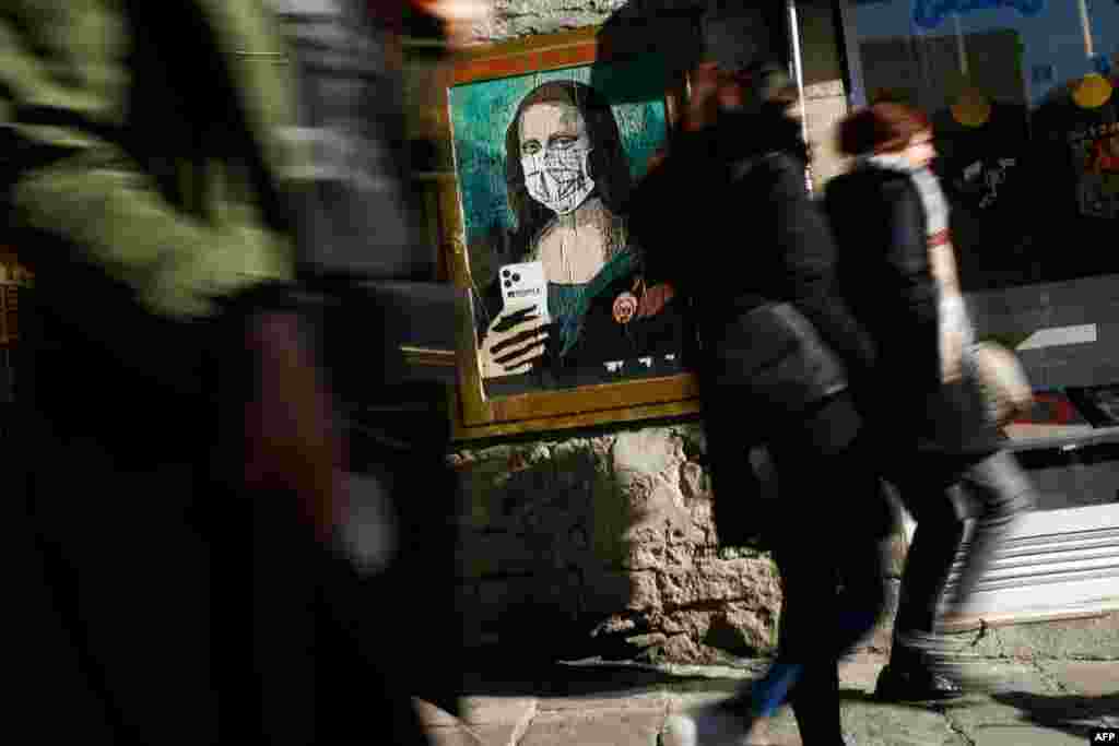 People walk by a sign by Italian urban artist Salvatore Benintende ( also known as &quot;TVBOY&quot;) showing Leonardo da Vinci&#39;s Mona Lisa wearing a protective face mask and holding a mobile phone reading &quot;Mobile World Virus&quot; in a street of Barcelona, Spain.