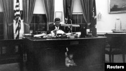 El presidente John F. Kennedy sentado en la oficina oval de la Casa Blanca mientras su hijo John F. Kennedy Jr. se esconde por debajo del escritorio.