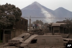 Rescue workers walk on rooftops in Escuintla, Guatemala, Monday, June 4, 2018, blanketed with heavy ash spewed by the Volcan de Fuego, or "Volcano of Fire," pictured in the background, left center. (AP Photo/Luis Soto)