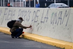 Seorang pengunjuk rasa bertopeng menuliskan grafiti di bawah pesan yang mengatakan dalam bahasa Spanyol "Porros keluar dari UNAM," di luar Universitas Otonomi Nasional Meksiko, UNAM. (Foto: AP/Rebecca Blackwell)