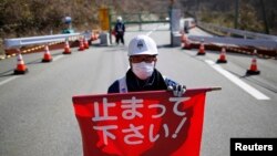 Nhân viên an ninh cầm cờ đỏ 'Xin dừng lại!' trước cổng vào thị trấn Tamura và Okuma ở Fukushima. Hơn 120.000 cư dân sống trong khu vực có bán kính 20km bao quanh Nhà máy điện hạt nhân Daiichi ở Fukushima vẫn chưa trở về lại nhà cửa của họ được vì mức phóng xạ ở đó vẫn còn cao.