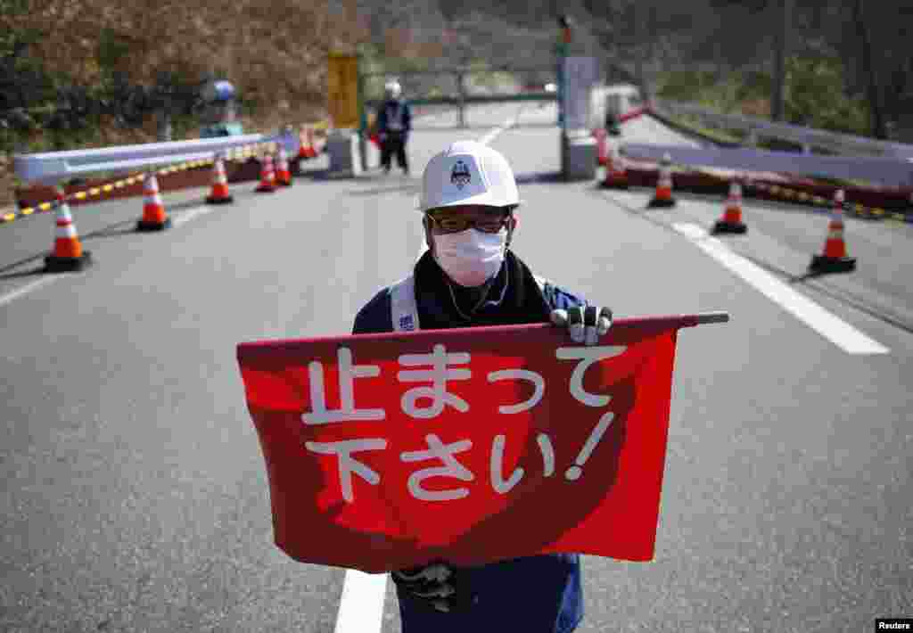 A security guard holds a flag that reads &quot;Please Stop!&quot;, as he stands by a steel gate that marks the border between Tamura and Okuma town in Okuma town, Fukushima prefecture, April 1, 2014.