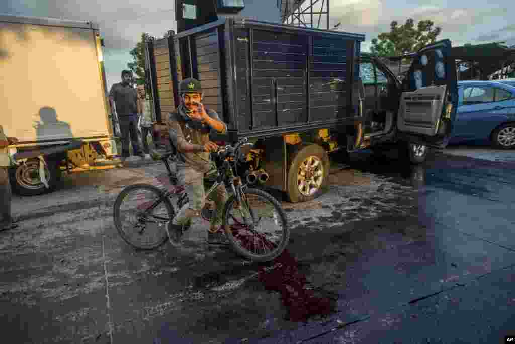 Un hombre toma en su mano un cartucho de bala en una calle ensangrentada. A su lado se ve un camión con una rueda pinchada, cubierto de balazos.