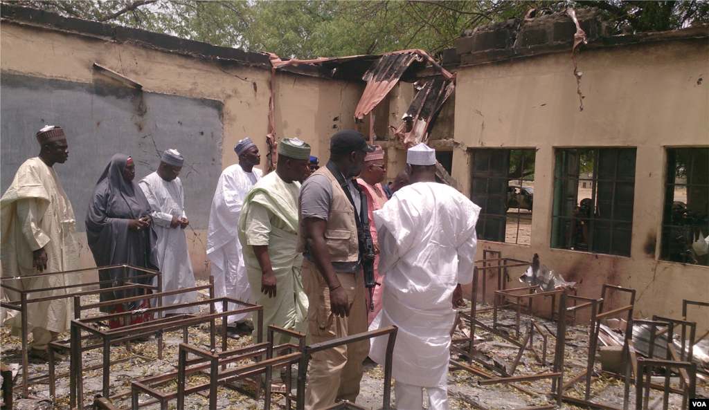 The governor of Chibok (in rose-colored clothing), the local education commissioner (fourth from left) with armed security and townspeople inspect the government secondary school that was set on fire after the more than 200 girls were abducted by gunmen, Chibok, Niger, April 21, 2014. (Anne Look/VOA)