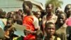 Refugees stand with a placard that reads: #ImmediateRelocation. Many refugees want to go to a settlement camp in Adjumani, but officials say these are full. ( Photo: Lizabeth Paulat for VOA)