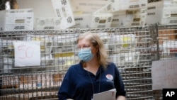 FILE- Cages loaded with ballots in U.S. Postal Service bins rest behind a worker at a Board of Elections facility in New York, July, 22, 2020. 