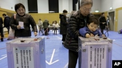 ເດັກຊາຍຜູ້ນຶ່ງເອົາບັດລົງຄະແນນສຽງຂອງແມ່ຕູ້ລາວ ໃສ່ໃນກ່ອງເກັບບັດເລືອກຕັ້ງ ຢູ່ສະຖານທີ່ປ່ອນບັດແຫ່ງນຶ່ງ ໃນນະຄອນໂຕກຽວ, ວັນອາທິດ ທີ 16 ທັນວາ 2012. 