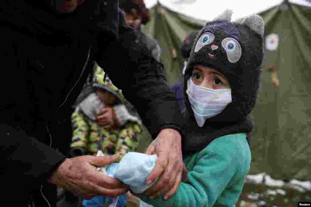 Seorang perempuan menutupi tangan seorang anak perempuan migran dengan masker wajah agar tetap hangat selama hujan salju di perbatasan Belarusia-Polandia, di wilayah Grodno, Belarus. (Foto: Reuters)