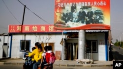 FILE - A Uyghur woman fetches schoolchildren as they ride past a picture showing China's President Xi Jinping joining hands with a group of Uyghur elders at the Unity New Village in Hotan, in western China's Xinjiang region, Sept. 20, 2018.