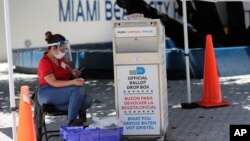 A poll worker wears personal protective equipment as she monitors a ballot drop box for mail-in ballots outside of a polling station during early voting, in Miami Beach, Florida. Aug. 7, 2020.