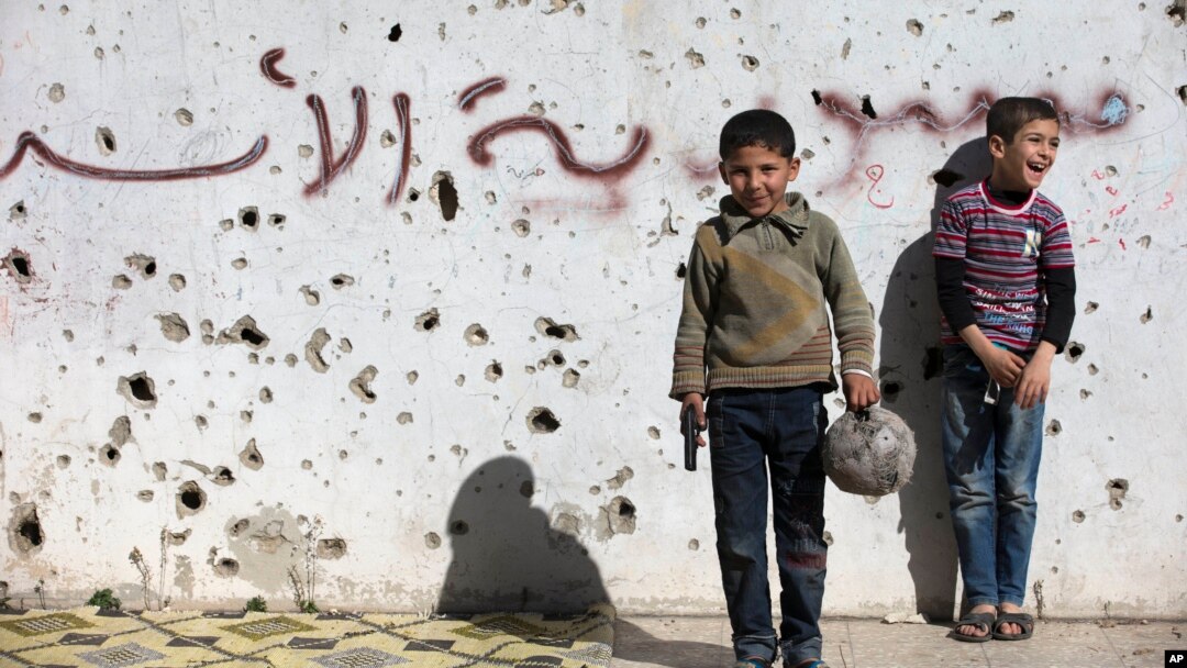 Un niño sirio sostiene una pistola de juguete mientras juega al fútbol con otros entre edificios destruidos en lo que un graffiti dice "Siria al-Assad" en la ciudad vieja de Homs, Siria, el viernes, 26 de febrero de 2016.