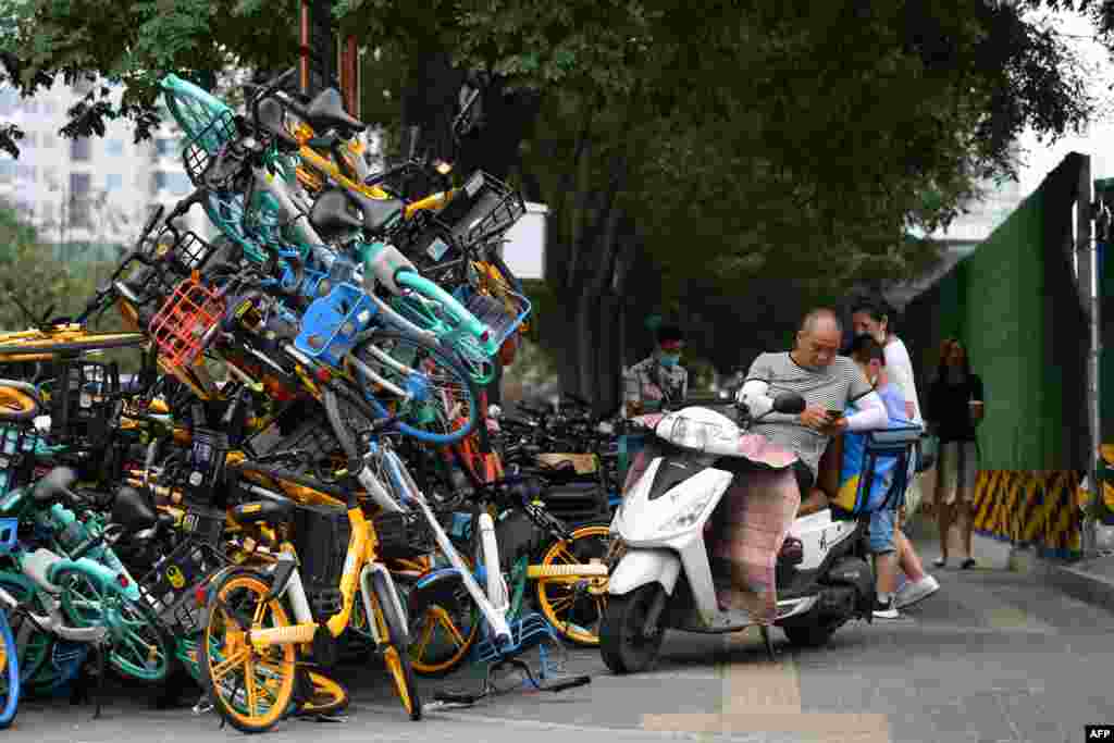 A delivery rider rests on a scooter next to a stack of shared bicycles on a sidewalk in Beijing, China.