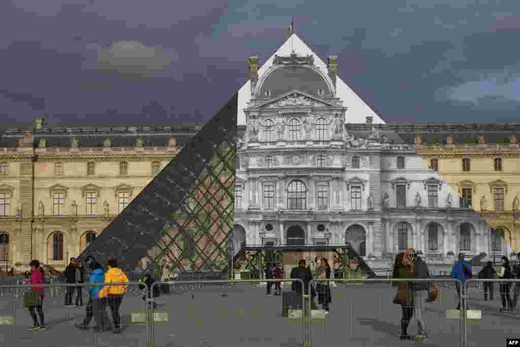 Para wisatawan mengunjungi Piramida Louvre di Paris, yang ditutup oleh gambar foto museum Louvre oleh seniman Perancis JR.