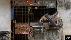 Un niño observa desde detrás de las rejas de una tienda de electrónica abierta mientras un soldado hace guardia en un punto de control de seguridad colocado por el ejército en Durán, Ecuador, el lunes 14 de agosto de 2023. [AP Photo/Martin Mejia]