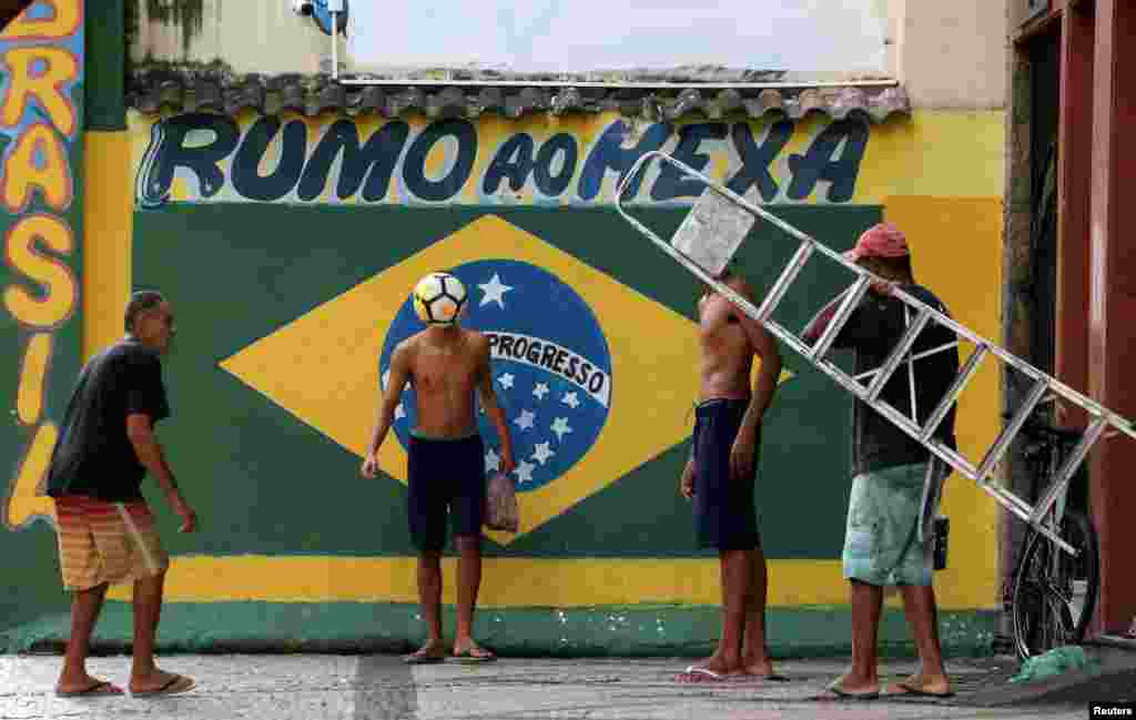 Beberapa warga bermain sepak bola di dekat mural bendera Brazil menjelang Piala Dunia 2018 di kawasan Gloria, Rio de Janeiro, Brazil, 12 Juni 2018. Pada mural tertera tulisan &quot;Mengejar Gelar Keenam.&quot; (Foto: Reuters)