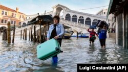 In this Monday, Dec. 23, 2019 file photo, people carry their belongings as they move through water during a high tide of 1.44 meters, near the Rialto Bridge, in Venice, Italy. (AP Photo/Luigi Costantini, file)