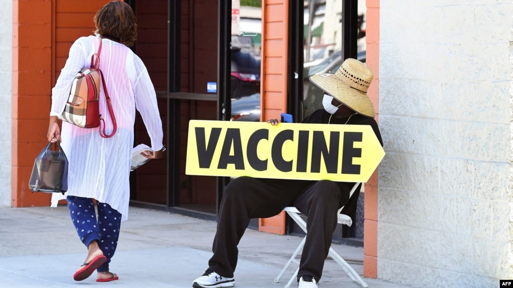 在加州洛杉矶，一位坐在椅子上的戴口罩男士端着一个指向疫苗接种诊所的方向标识牌。(2021年7月16日)(photo:VOA)