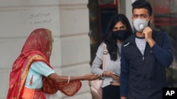 An elderly Indian woman seeks alms as youth wearing pollution masks walk through a shopping area in New Delhi, India, Nov. 14, 2019.