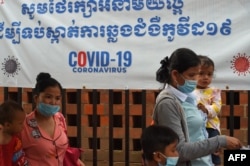 FILE - Women wearing face masks amid the coronavirus pandemic walk with their children outside a hospital with a banner warning about the dangers of the virus, in Phnom Penh, Cambodia, Sept. 29, 2020.