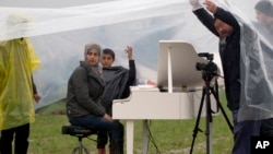 Syrian pianist Nour Alkhzam, center left, sits at the piano as Chinese dissident artist Ai Weiwei, right, holds a plastic sheet, during a heavy rain at the northern Greek border station of Idomeni, March 12, 2016.