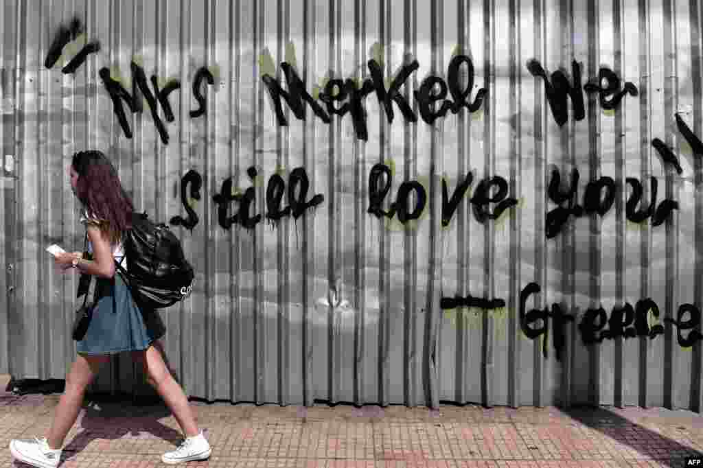 A young woman walks past graffiti reading &quot;Mrs. Merkel we still love you - Greece&quot; in central Athens. The European Central Bank&#39;s decision-making governing council held an emergency session Friday to discuss a request for an increase in liquidity to Greek banks.