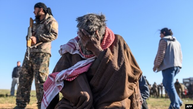 A member of the Kurdish-led Syrian Democratic Forces (SDF) stands by, left, as a man sits with his head in his hand, after leaving the Islamic State (IS) group's last holdout of Baghuz, in the eastern Syria, March 1, 2019.