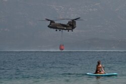 A Ch-47D Chinook helicopter is watched by a paddleboarder as it fills up with water while firefighting near Lambiri Beach at Patras on Aug. 1, 2021.