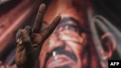 FILE - A Sudanese protester flashes a victory sign in front of a banner depicting ousted and detained president Omar al-Bashir, during a protest outside the army headquarters in the capital Khartoum, April 19, 2019.