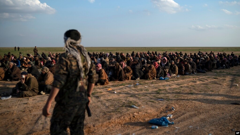 Kombatan Pasukan Demokratik Suriah yang didukung AS, berjaga di dekat para pria yang menunggu untuk diperiksa setelah dievakuasi dari wilayah terakhir ISIS, dekat Baghuz, timur Suriah, 22 Februari 2019. 