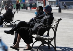 A couple wear face masks as they sit on a bench in the sun April 2, 2020, at the Coney Island boardwalk in the Brooklyn borough of New York.