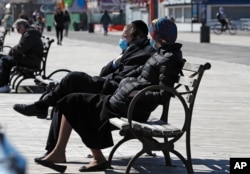 A couple wear face masks as they sit on a bench in the sun April 2, 2020, at the Coney Island boardwalk in the Brooklyn borough of New York.