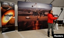 ຜູ້ທ່ຽວຊົມຄົນນຶ່ງ ກຳລັງບັນທຶກພາບຂອງຕົນເອງ ໃນຂະນະຢືນຢູ່ຕໍ່ໜ້າປ້າຍວາງສະແດງ ຂອງຍານອະວະກາດ ສຳຫຼວດດາວພະອັງຄານ ຂອງອົງການ NASA ຢູ່ໃນສະໜາມບິນ Lompoc ກ່ອນການ ຍິງຂຶ້ນສູ່ ອະວະກາດດ້ວຍຈະຫຼວດ ລຸ້ນ V ຂອງບໍລິສັດ United Launch Alliance Atlas ທີ່ຈະບັນທຸກ ຍານອະວະກາດສຳຫຼວດ InSight Mars lander ຂອງອົງການ NASA ຊຶ່ງໄດ້ຖະຍານຂຶ້ນຈາກ ຖານທັບອາກາດ Vandenberg ໃນລັດແຄລີຟໍເນຍ, ວັນທີ 5 ພຶດສະພາ 2018.