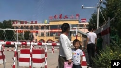 In this Aug. 31, 2018, file photo, a child and a woman wait outside a school entrance mounted with surveillance cameras and barricades with multiple layers of barbed wire in Peyzawat, western China's Xinjiang region. (AP Photo/Ng Han Guan, File)